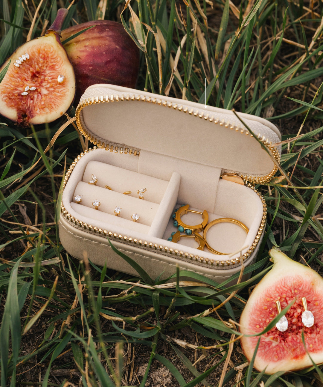An open cream-colored jewelry case with gold trim sits on green grass outdoors, surrounded by sliced figs. The case contains several gold rings and a small gold tool, and the jewelry pieces are shown close up.