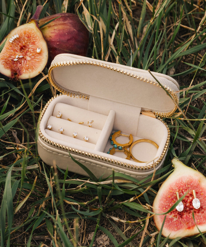 An open cream-colored jewelry case with gold trim sits on green grass outdoors, surrounded by sliced figs. The case contains several gold rings and a small gold tool, and the jewelry pieces are shown close up.